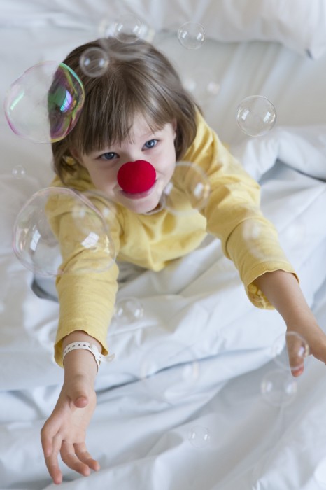 Ein Mädchen mit roter Schaumstoffnase schaut lachend in die Kamera. Sie sitzt im Krankenbett, um sie herum fliegen Seifenblasen durch die Luft.