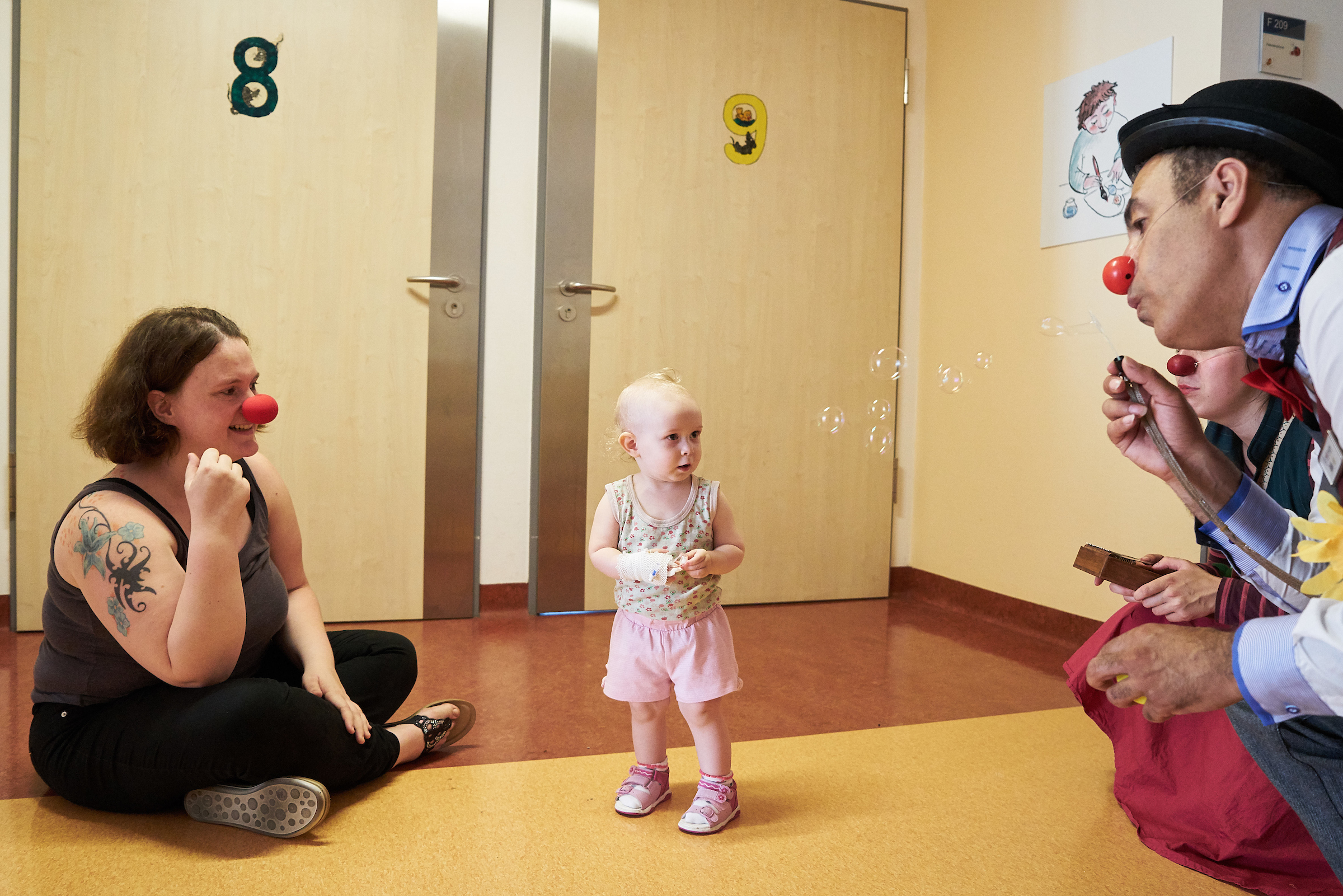 Zwei Clowns hocken vor einem kleinem Kind mit seiner Mutter und es staunt über die Seifenblasen 