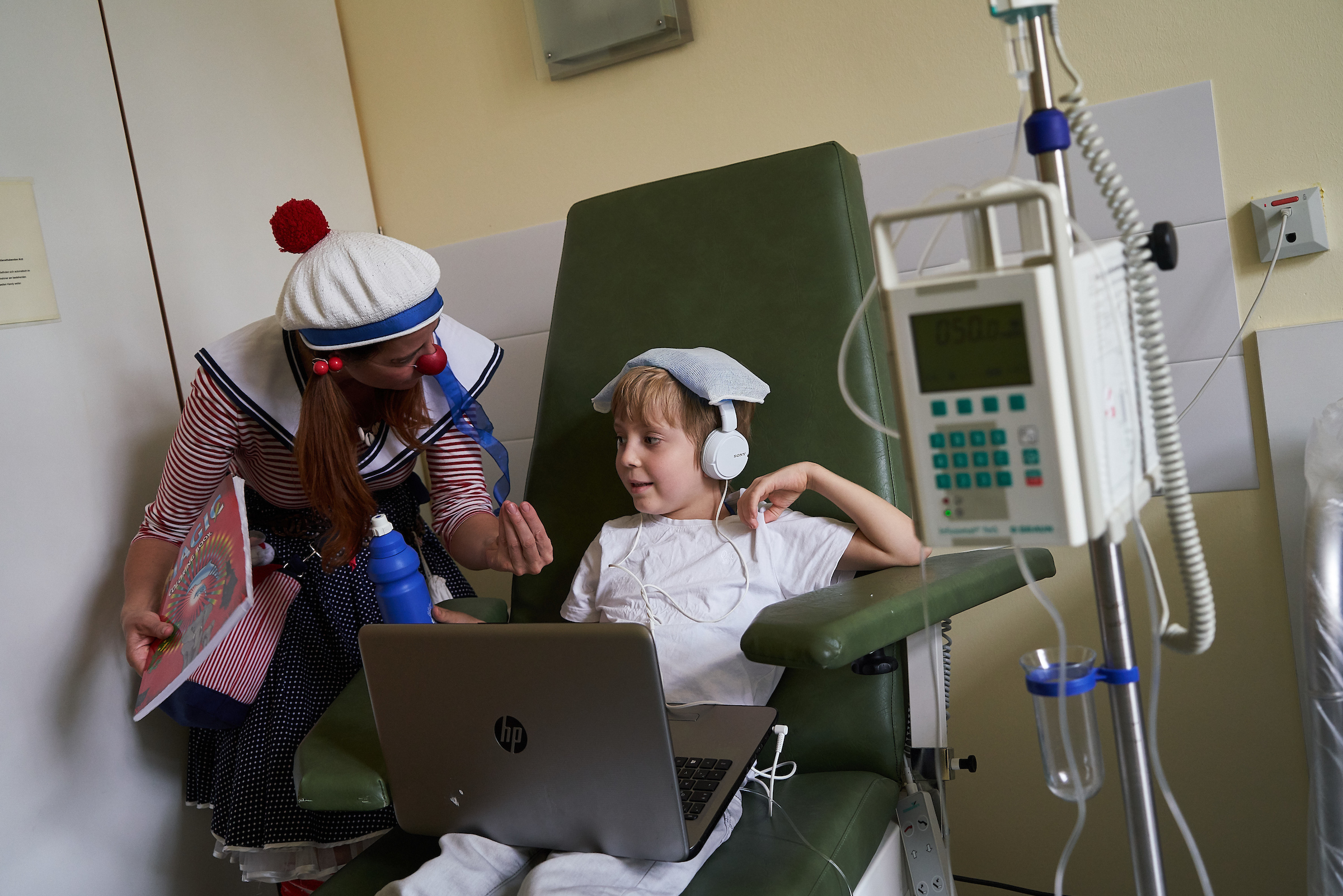 Ein Clown steht neben einem Kind was auf einem Stuhl vor einem Leptop sietzt 