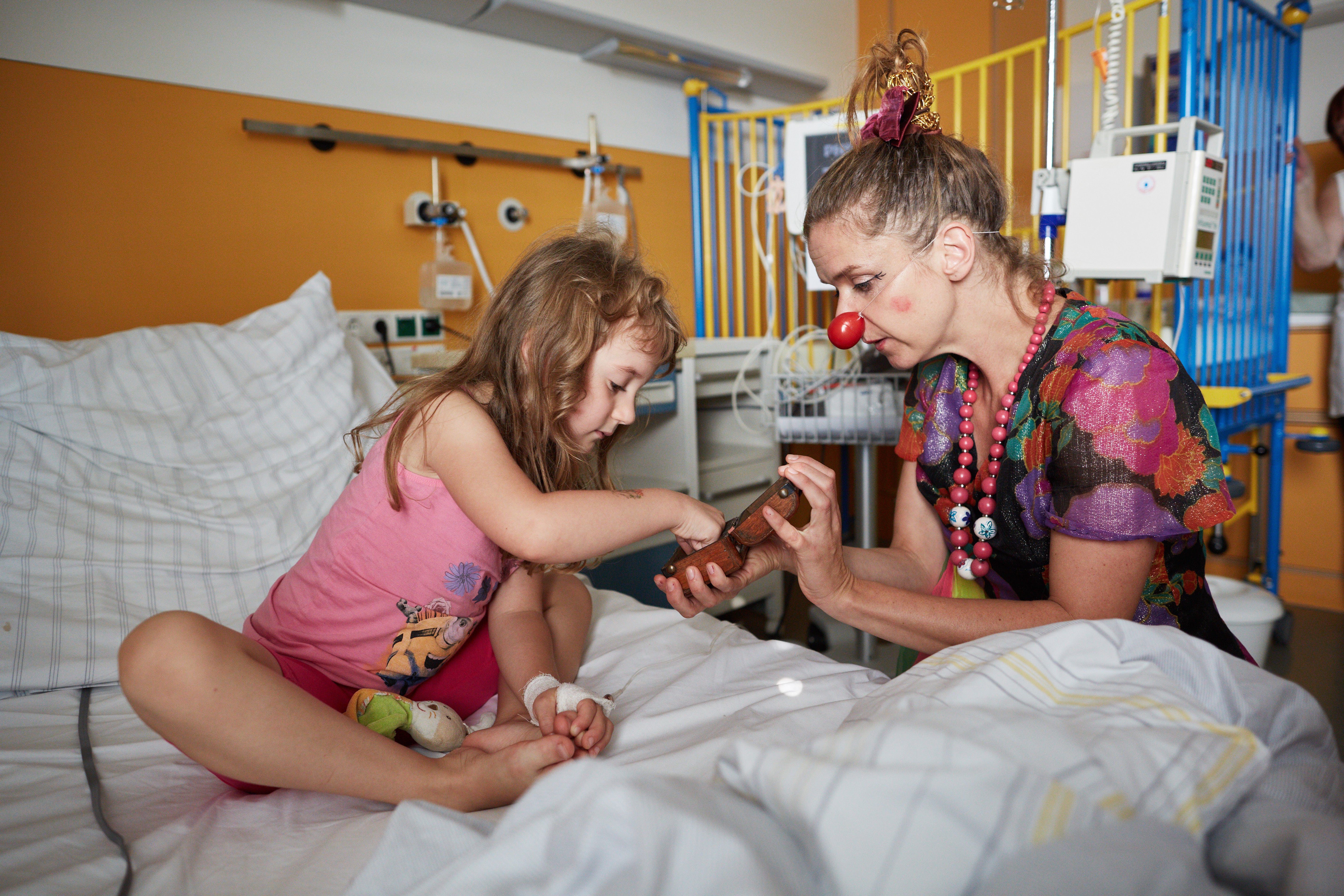 ein weiblicher Klinikclown zeigt einem klienne Mädchen im Krankenbett eine kleine Dose