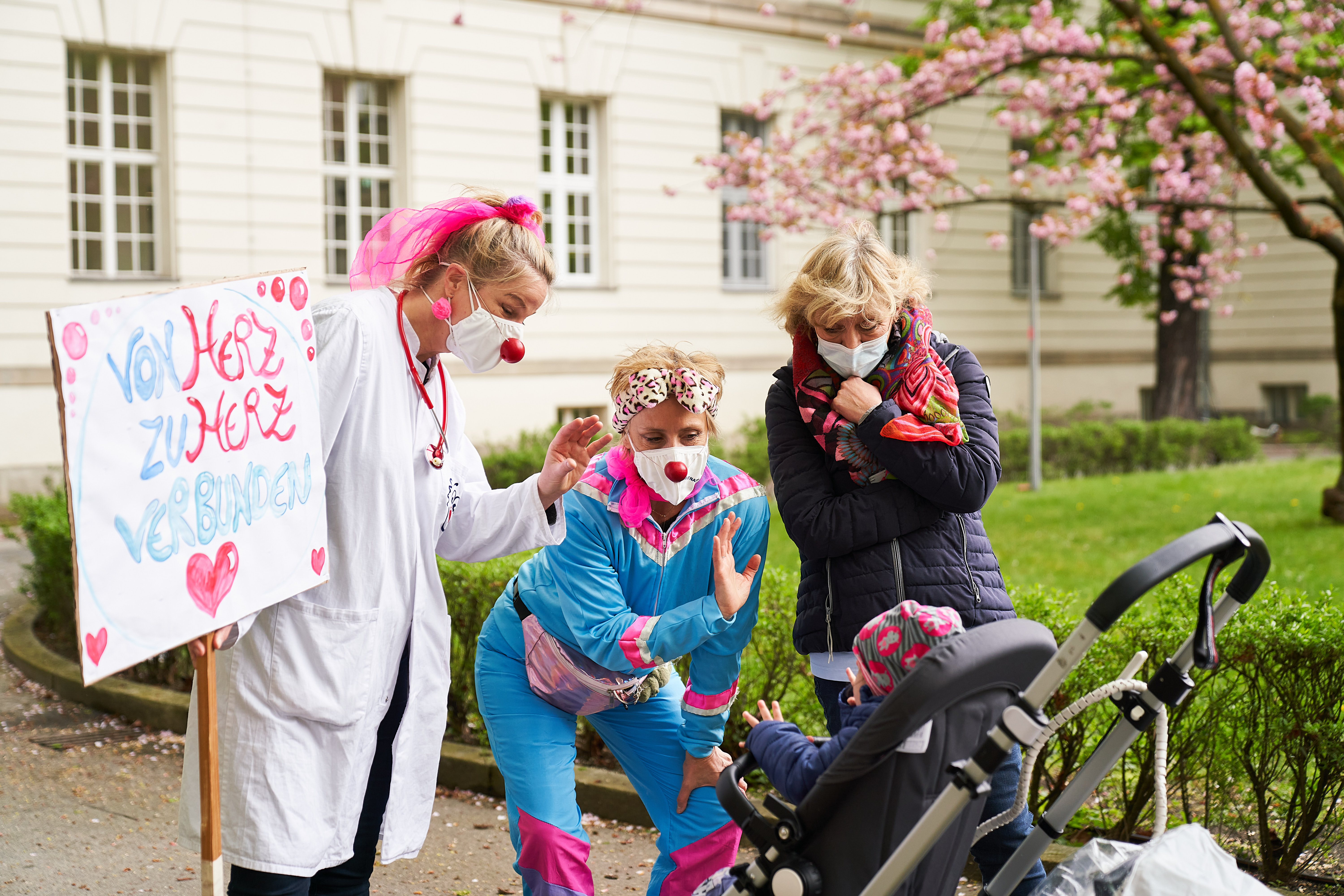 Zwei Klinikclowns von ROTE NASEN beugen sich lächelnd zu Kind im Kinderwagen hinunter, das an ein künstliches Herz angeschlossen ist