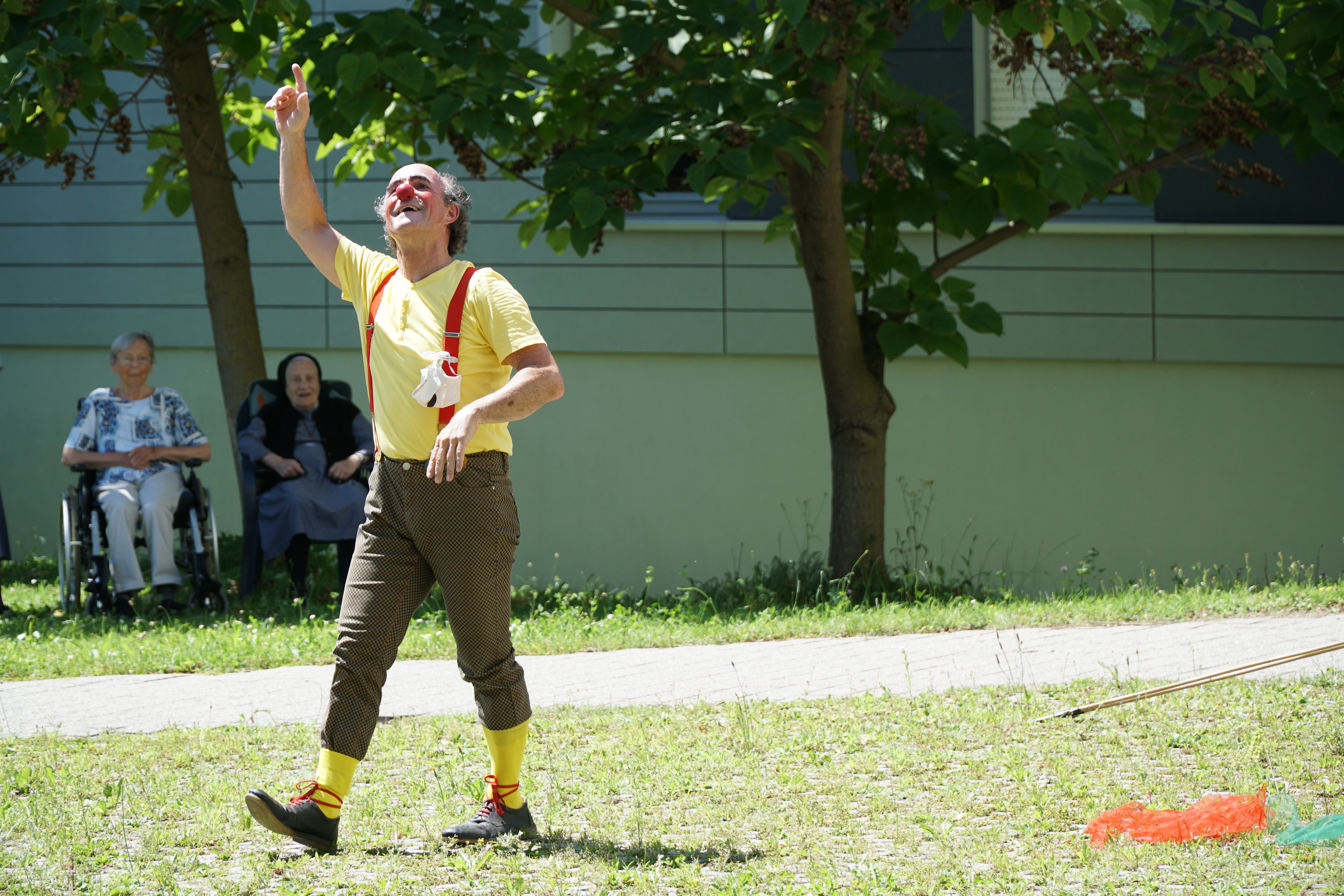 ein männlicher Clown winkt nach oben und zwei Damen sitzen im Schatten hinter ihm und schauen ihm zu