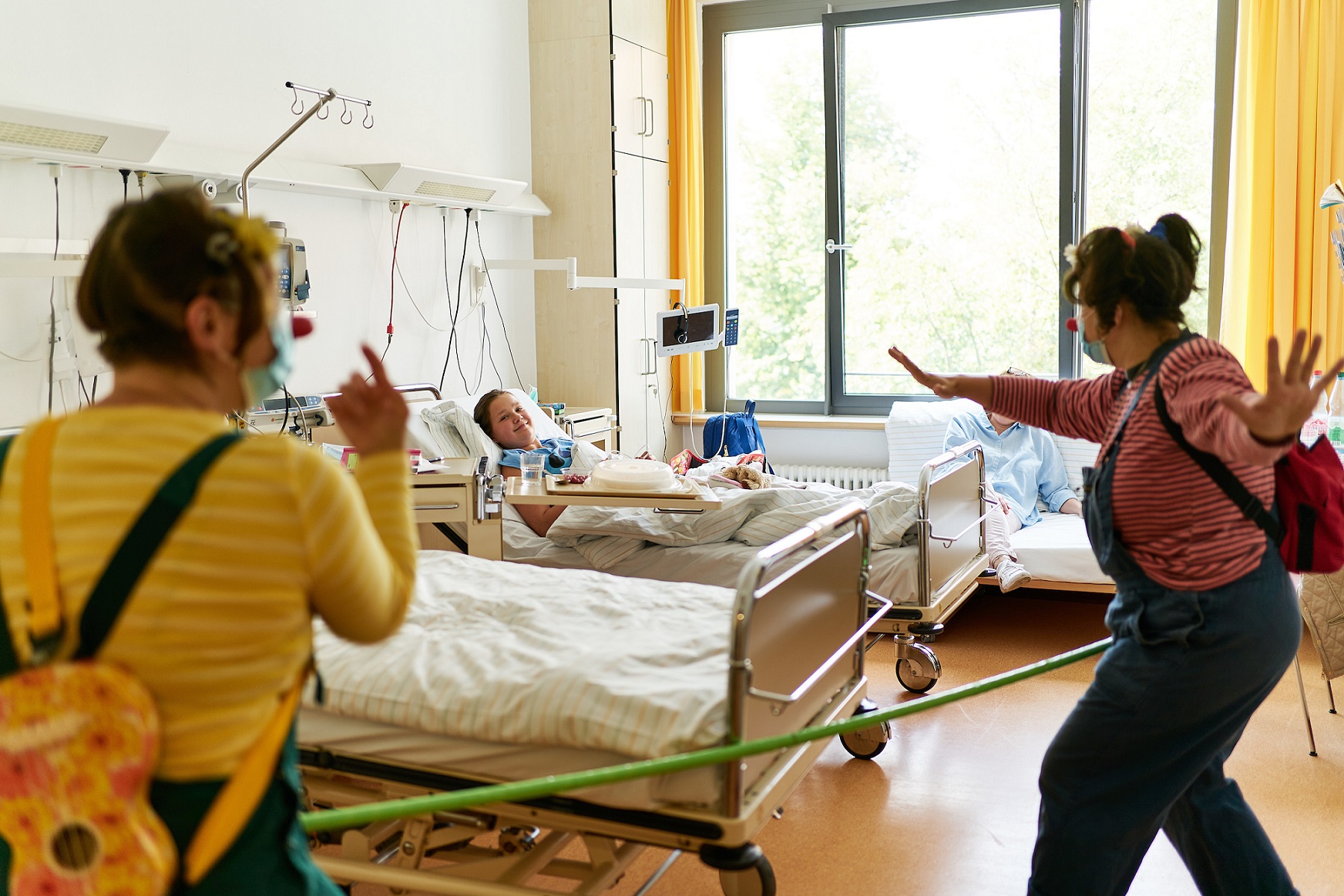 zwei ROTE NASEN Clowns mit Mund-Nasen-Schutz benutzen bei einer Clownvisite im Kinderkrankenhaus einen Meterstab als Abstandshalter. Corona.