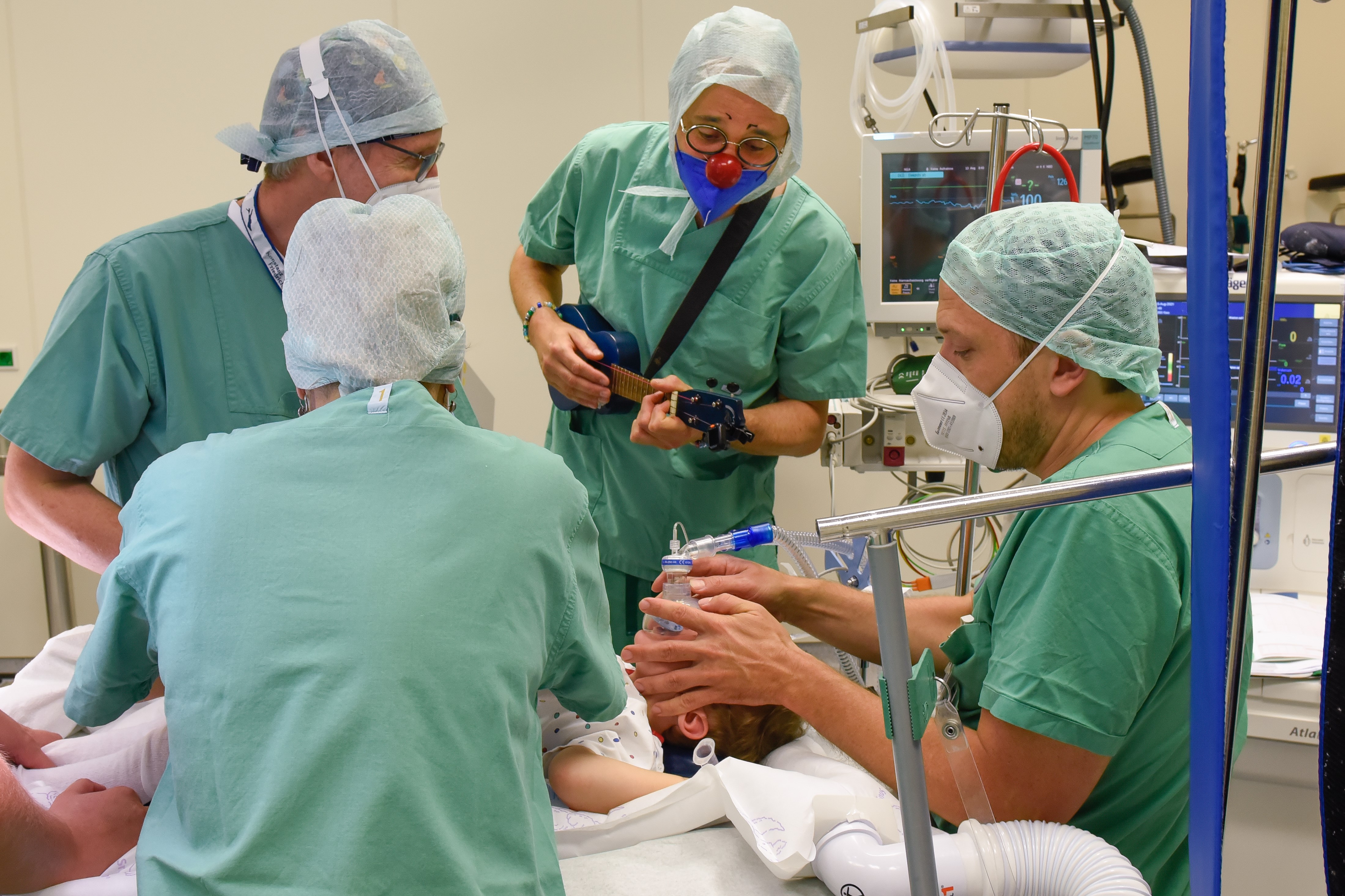 Im OP Saal stehen ein Anästhesist, ein OP Pfleger und ROTE NASEN Klinikclown Sören Kaspersinski am Bett eines kleinen Jungen in OP Hemdchen. Der Anästhesist narkotisiert den Jungen. Der Clown mit roter Nase und OP Kleidung spielt zur Beruhigung Ukulele. Das ist Teil der ROTE NASEN OP Begleitung im Potsdamer Klinikum Ernst von Bergmann.