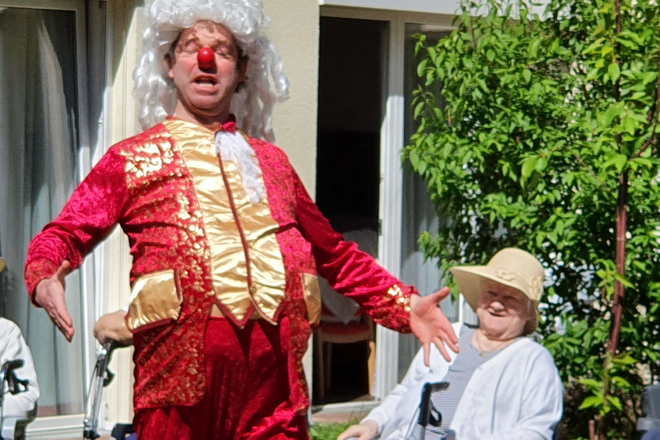 Rote Nasen Klinikclown Leopold Altenburg mit Barockperücke. Im Hintergrund lacht eine ältere Dame.