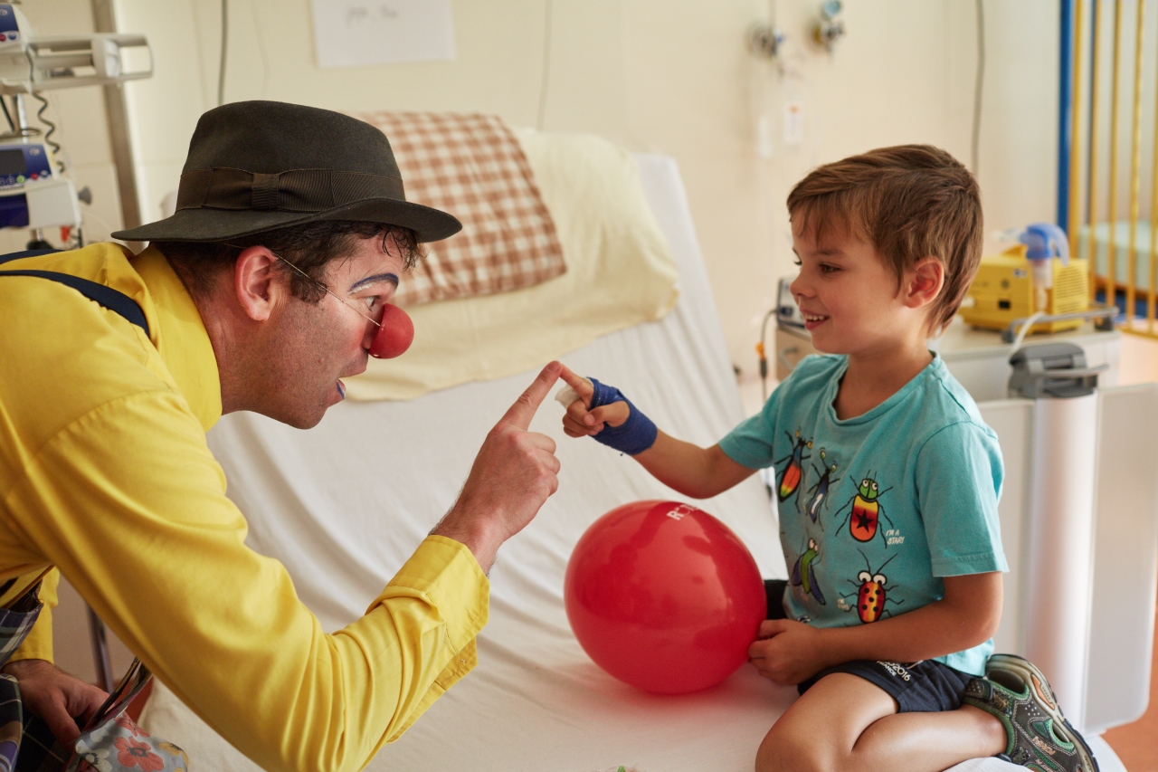  Der Klinikclown berührt den Zeigefinger eines kleines Jungen auf seinem Zimmer