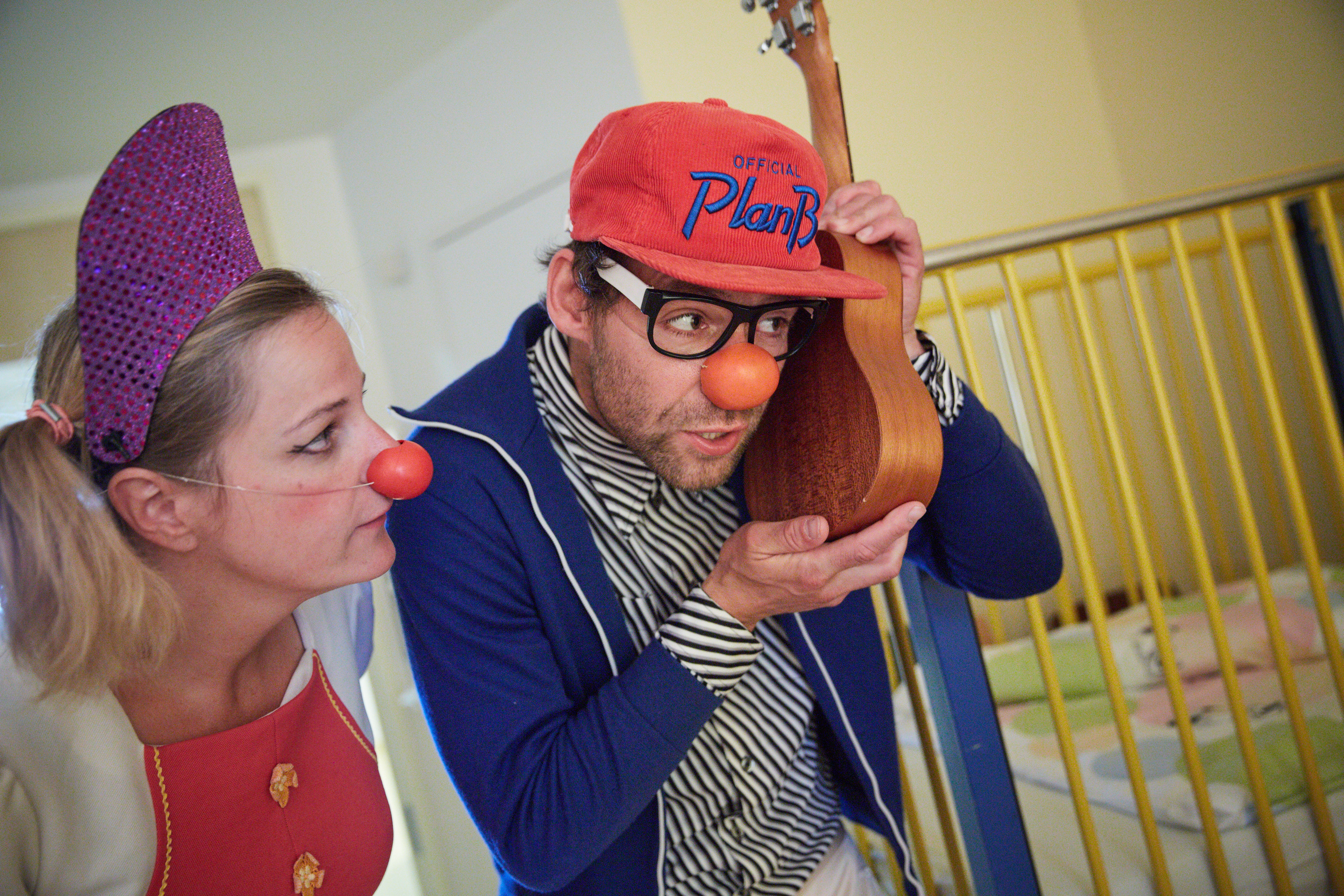 Ein Klinikclown hält sich eine Ukulele ans Ohr und seine Kollegin steht daneben