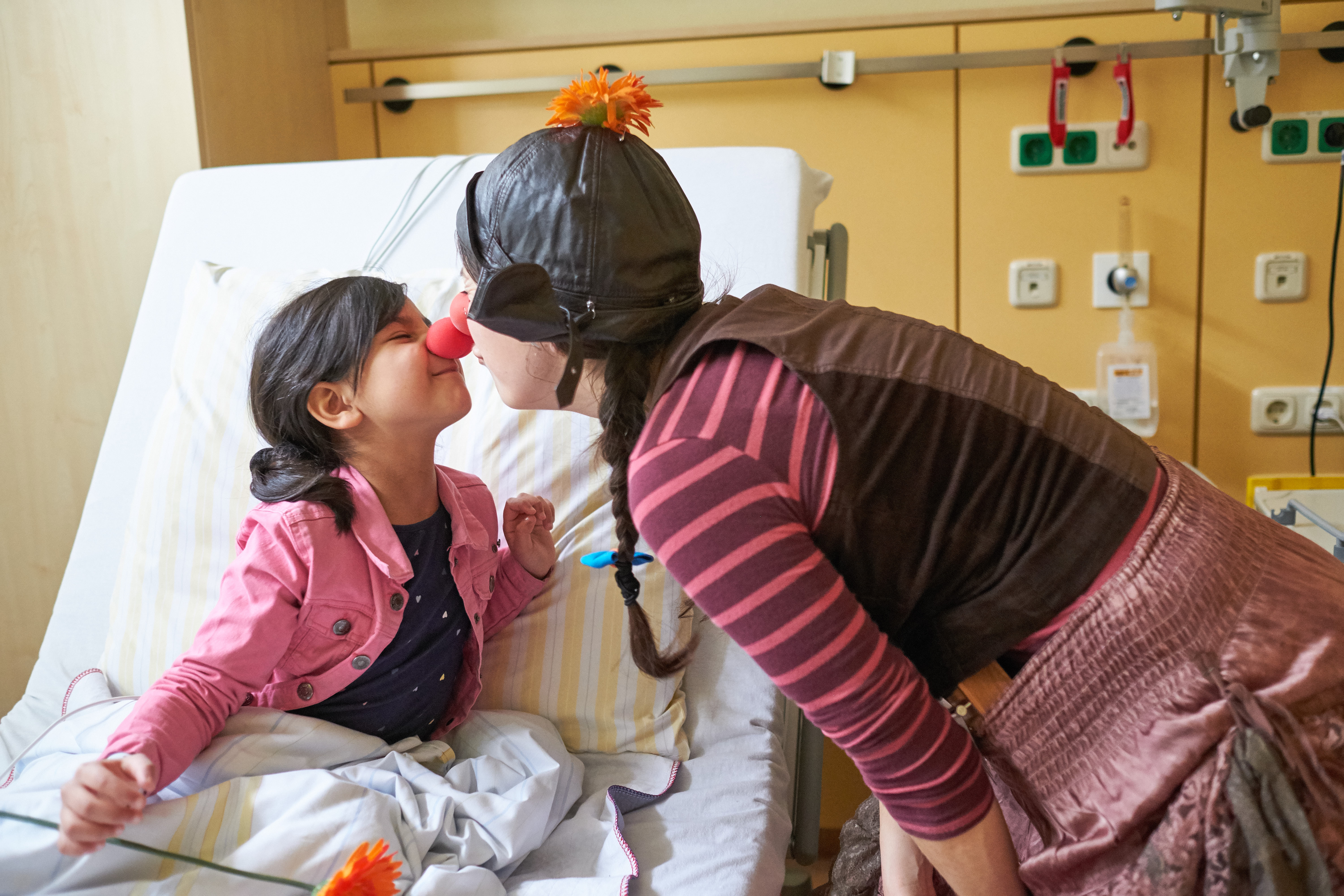 Ein Mädchen trägt in ihrem Krankenhausbett eine rote Schaumstoffnase und die Klinikclownin gibt ihr einen küss von Nase zu Nase